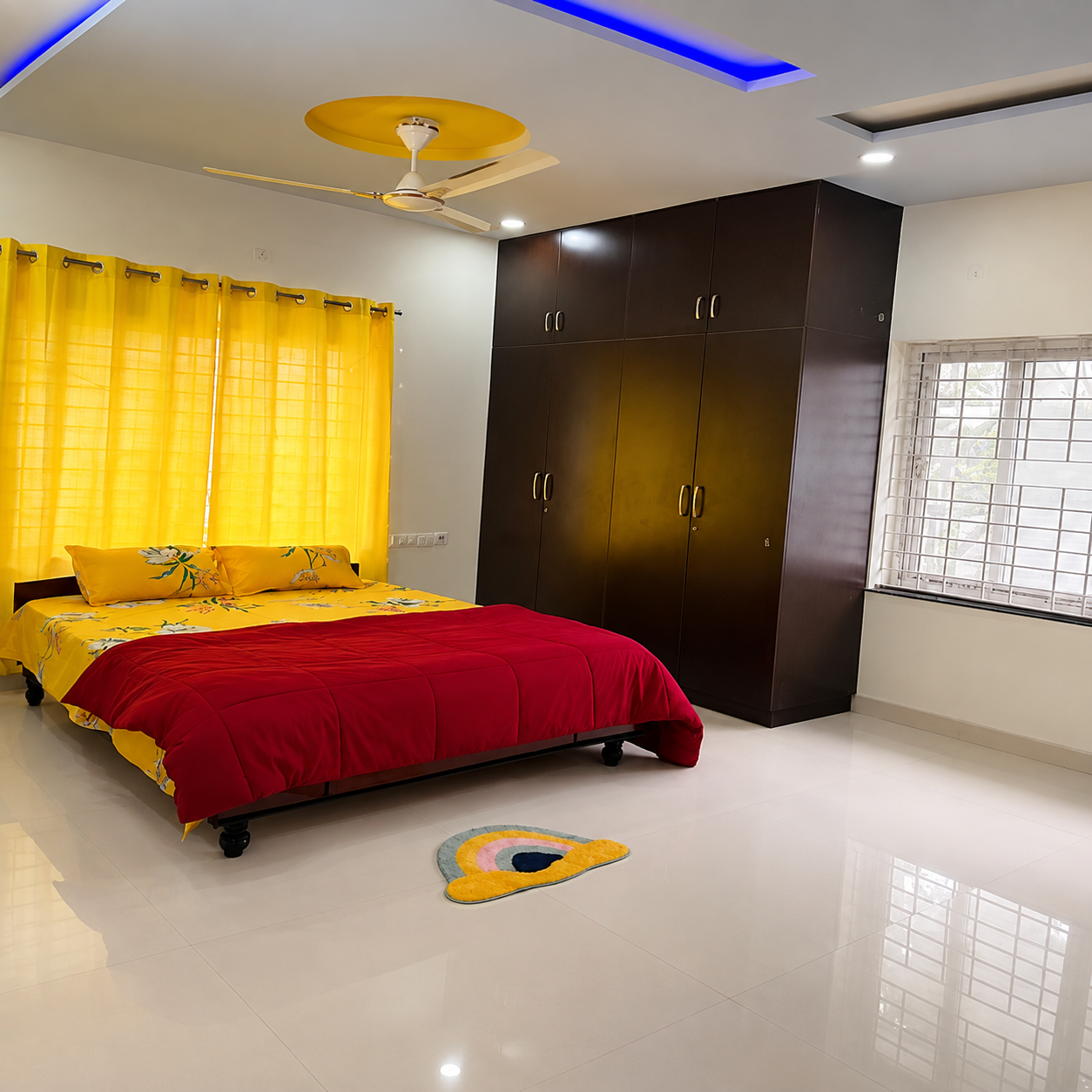 Bedroom, modern bedroom, spacious bedroom, contemporary bedroom interior, master bedroom, stylish bedroom, bright bedroom, well-lit bedroom, yellow bedroom decor, red bedspread, floral bedsheet, colorful bedroom, king-size bed, comfortable bed, bedroom furniture, dark wood wardrobe, large wardrobe, built-in wardrobe, ample storage, glossy floor, tiled bedroom floor, white walls, yellow curtains, window curtains, natural light, large window, ventilated bedroom, ceiling fan, false ceiling, LED ceiling lights, blue LED lighting, decorative ceiling, modern home interior, clean bedroom, organized bedroom, cozy bedroom, apartment bedroom, rental bedroom, student-friendly bedroom, student accommodation, student living space, study-friendly bedroom, business professional bedroom, professional accommodation, work-life balance space, relaxing bedroom, comfortable living space, urban home, practical bedroom design, affordable bedroom, residential interior, real estate bedroom.